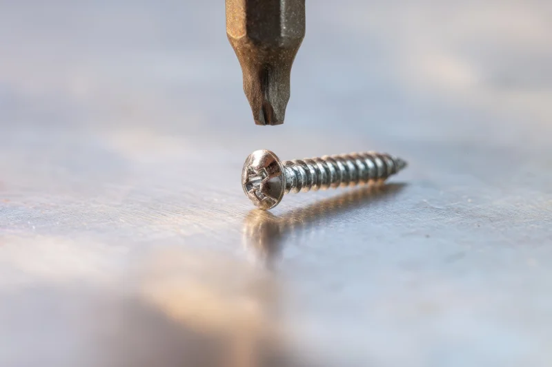 Metallic screw lying on wooden surface with screwdriver bit hovering above it