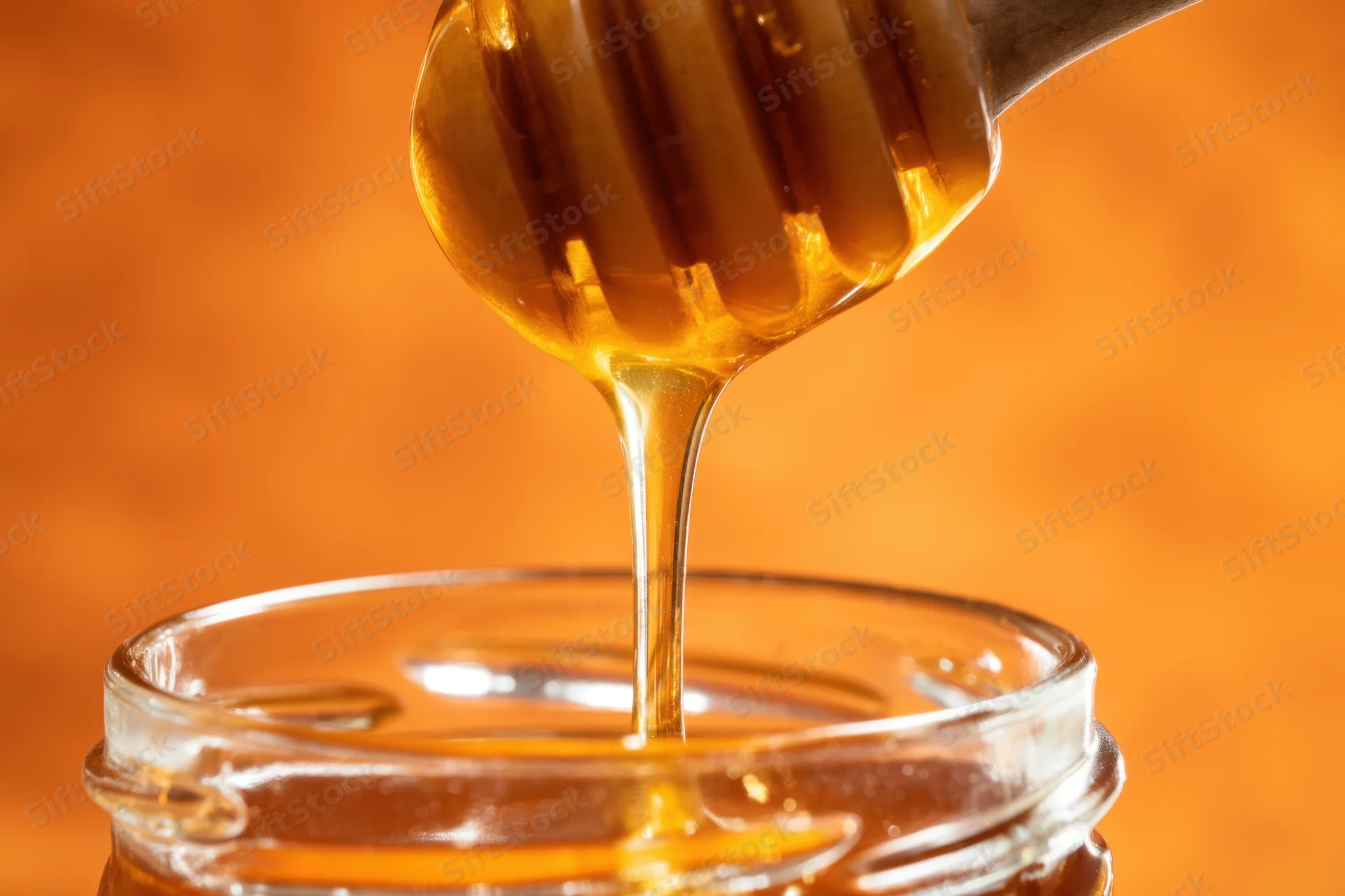 Golden honey dripping from wooden spoon into glass jar