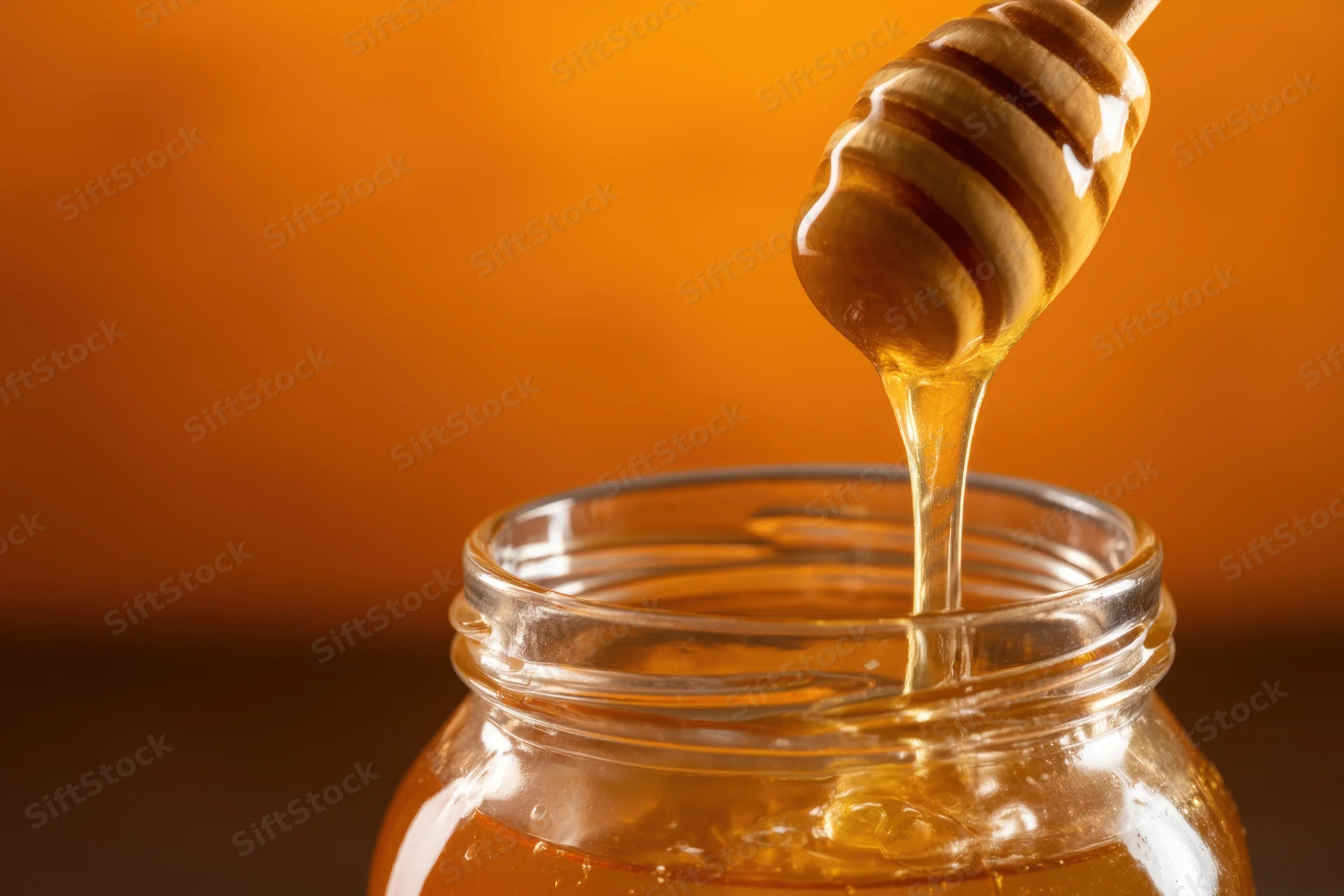 Golden honey dripping from a wooden dipper into a clear glass jar against a warm orange background