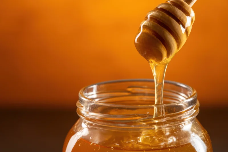 Golden honey dripping from a wooden dipper into a clear glass jar against a warm orange background