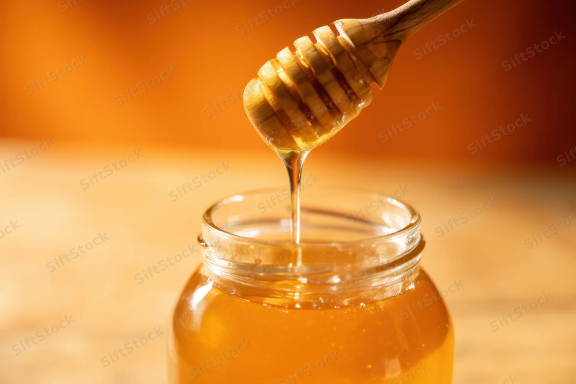 Golden honey dripping from wooden dipper into glass jar on wooden surface