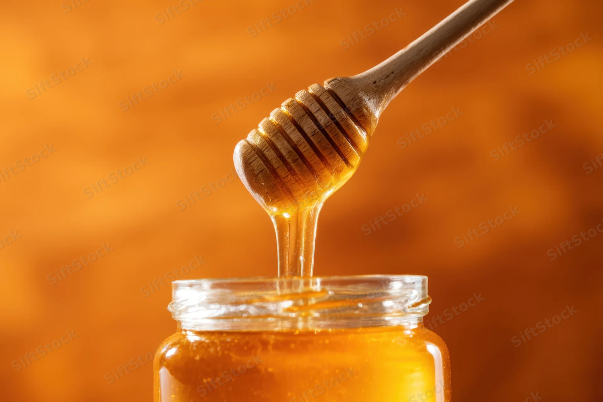 Golden honey dripping from wooden dipper into glass jar against warm background