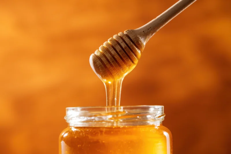 Golden honey dripping from wooden dipper into glass jar against warm background