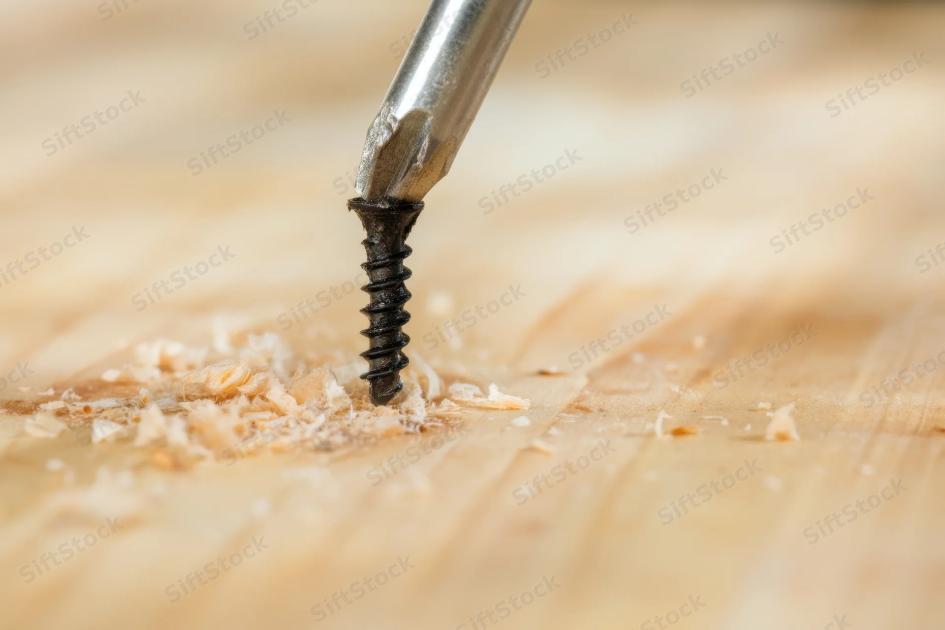 Metal screwdriver driving a black screw into wood, surrounded by wood shavings