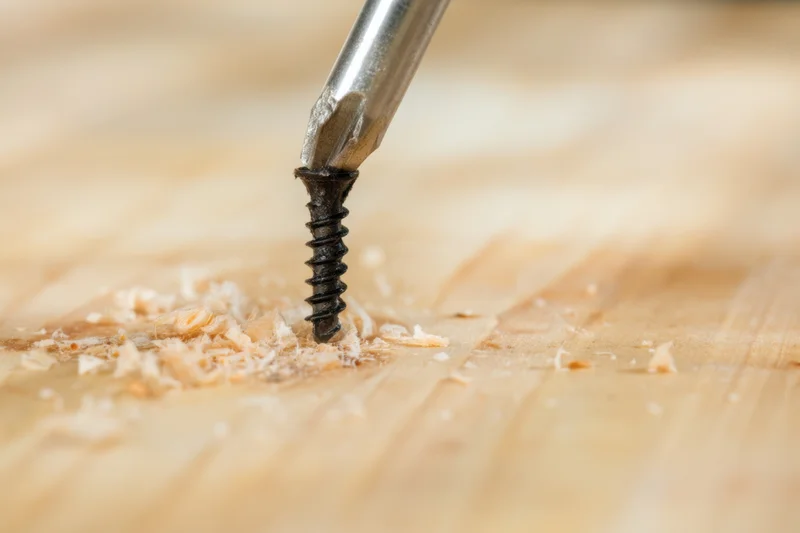 Metal screwdriver driving a black screw into wood, surrounded by wood shavings