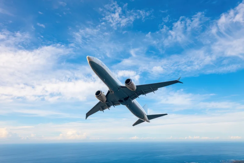 Commercial airplane ascending in flight over ocean under blue sky with scattered clouds
