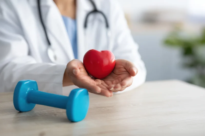 Doctor holding red heart shape with dumbbell on wooden table in clinic setting