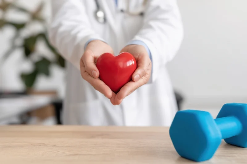 Doctor holding red heart shape in hands with blue dumbbell on wooden table in clinic setting