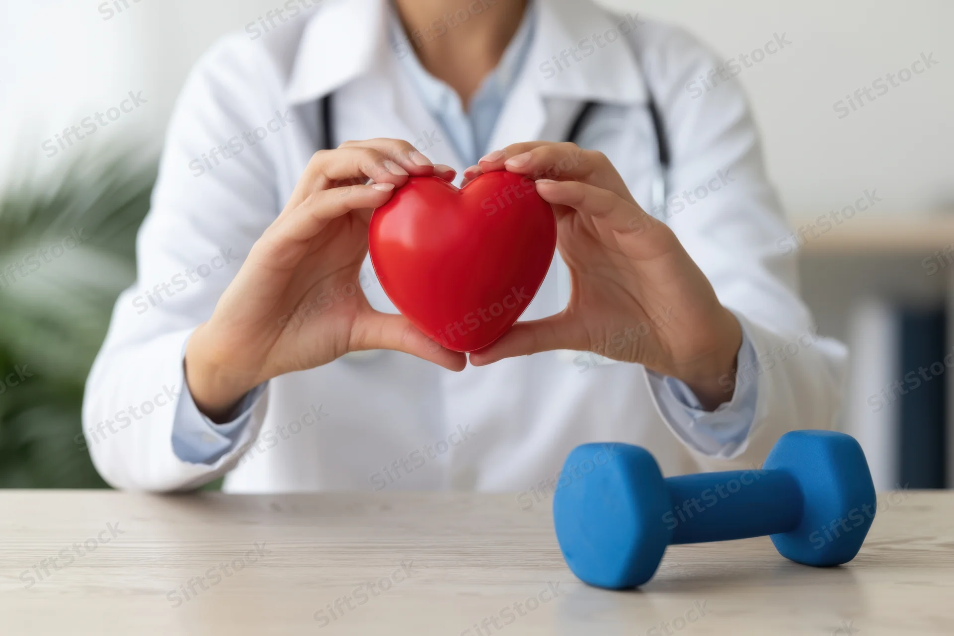 Doctor in white coat holding red heart shape with hands, blue dumbbell on wooden table in clinic setting