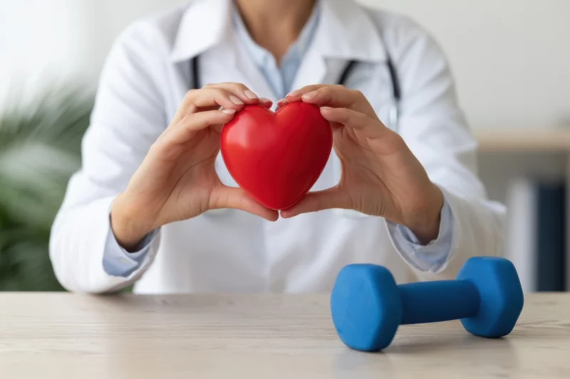 Doctor in white coat holding red heart shape with hands, blue dumbbell on wooden table in clinic setting