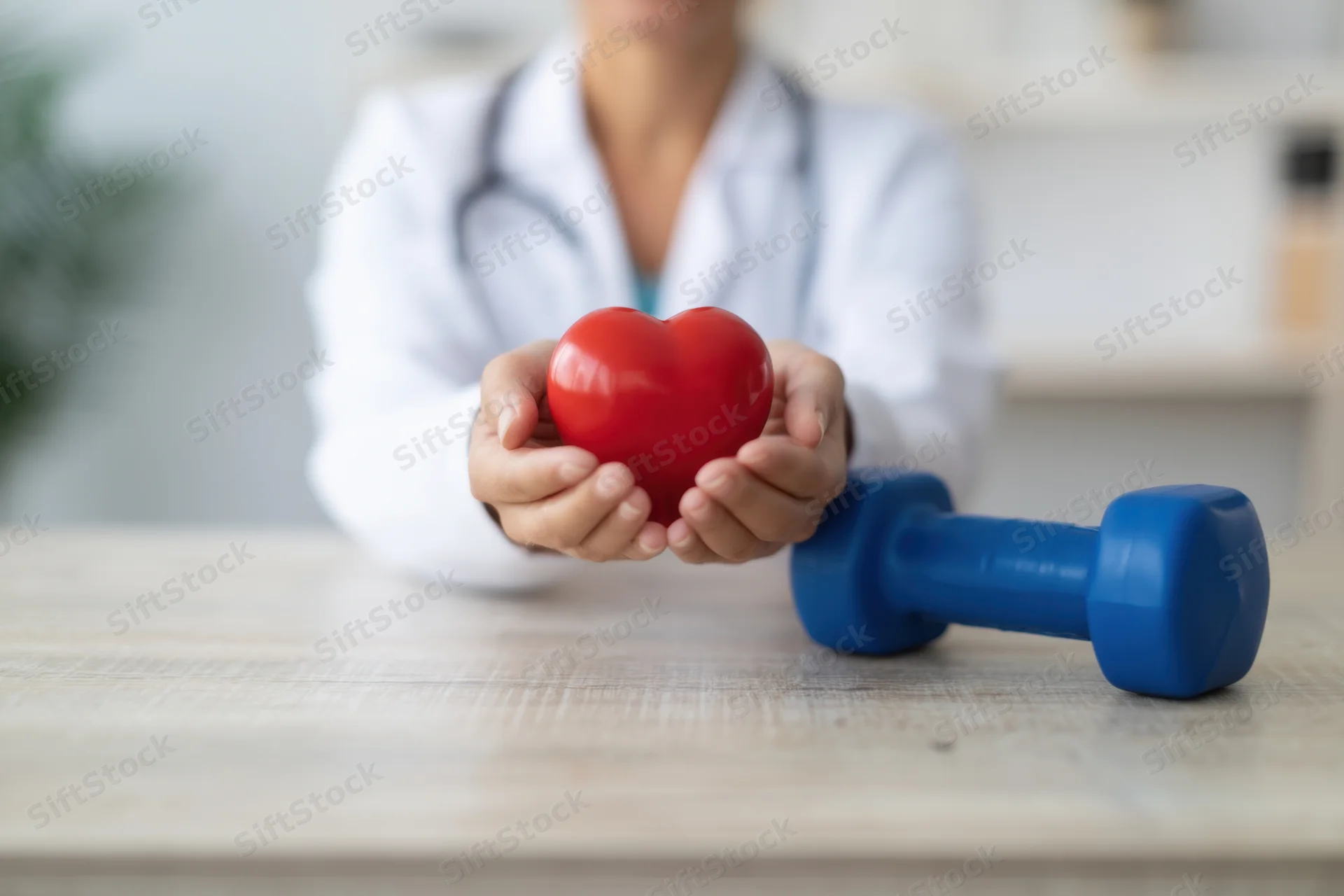 Doctor holding red heart shape on wooden table with blue dumbbell beside it
