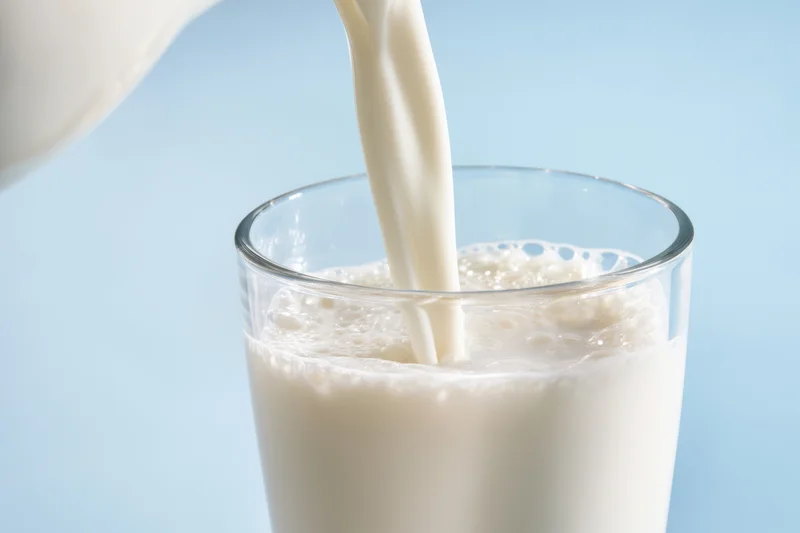 Milk being poured into a clear glass from a pitcher against a light blue background