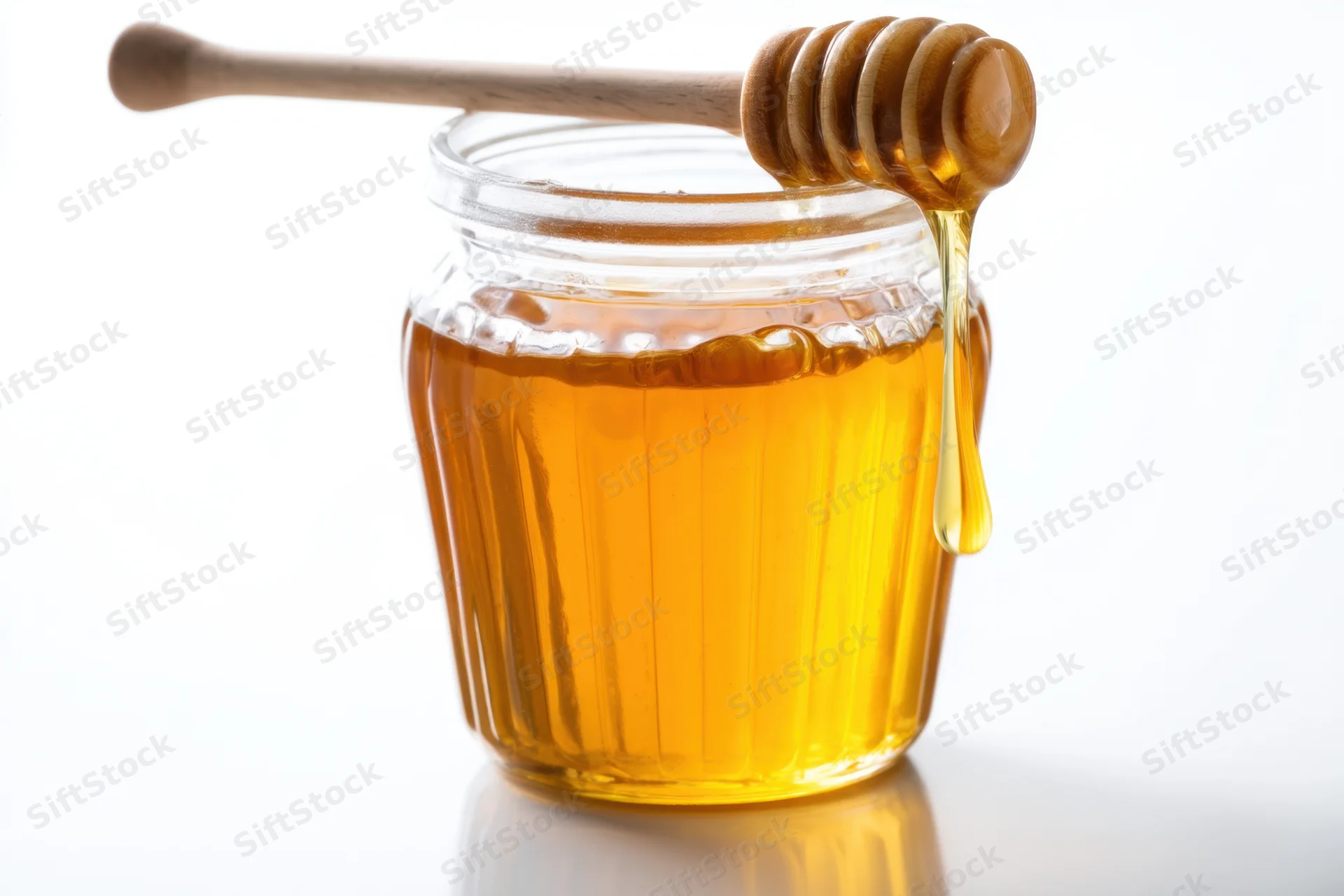 Golden honey dripping from wooden dipper into glass jar on white surface