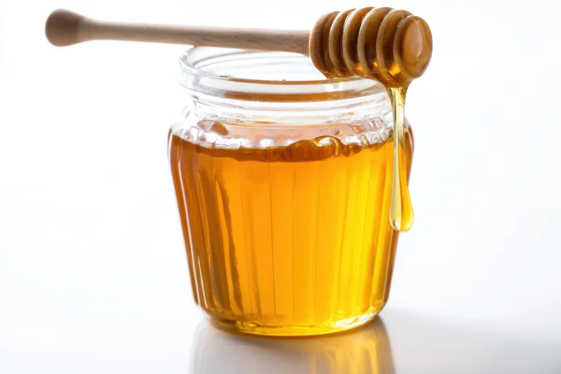 Golden honey dripping from wooden dipper into glass jar on white surface