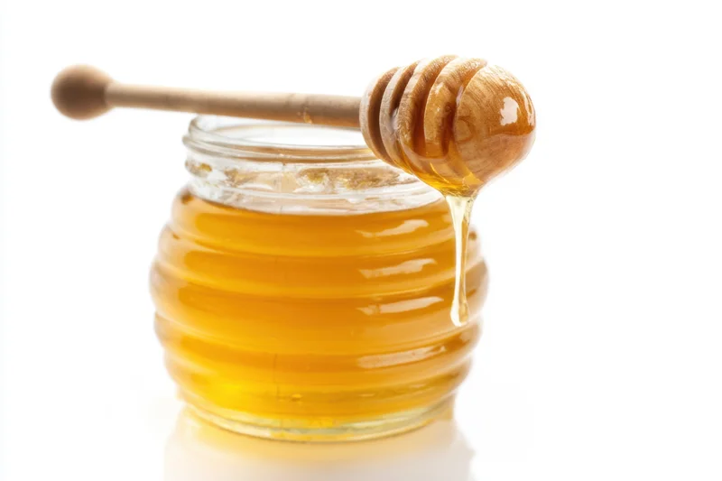 Golden honey dripping from wooden dipper into glass jar on white background