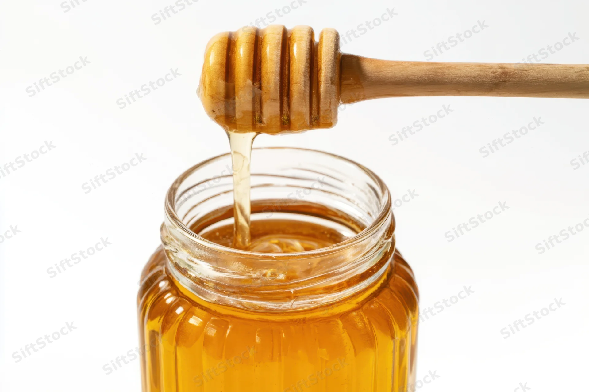 Golden honey dripping from wooden honey dipper into glass jar on white background