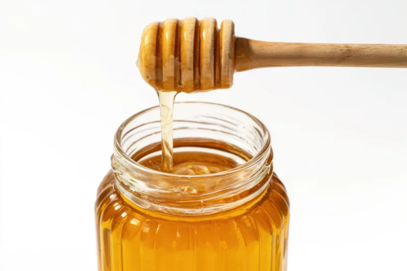 Golden honey dripping from wooden honey dipper into glass jar on white background