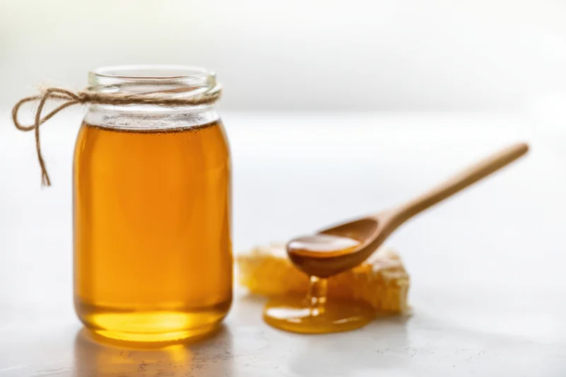 Glass jar of honey with wooden spoon and honeycomb on white surface
