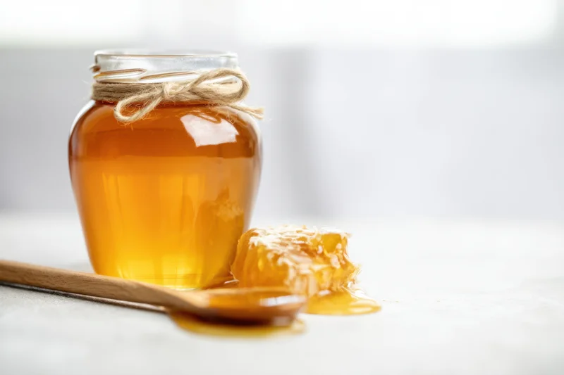 Glass jar of honey with twine tied around neck, wooden spoon and honeycomb beside it on light surface
