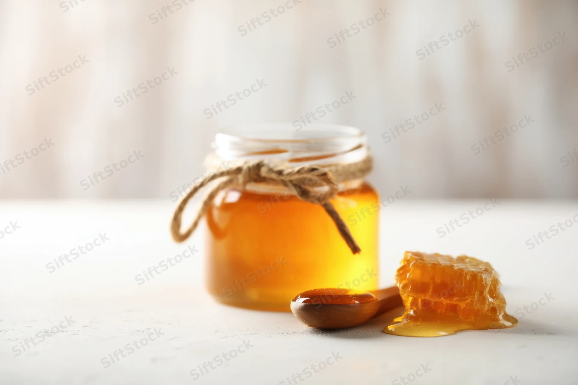 Glass jar of honey with twine tied around neck, wooden spoon and honeycomb beside it on white surface
