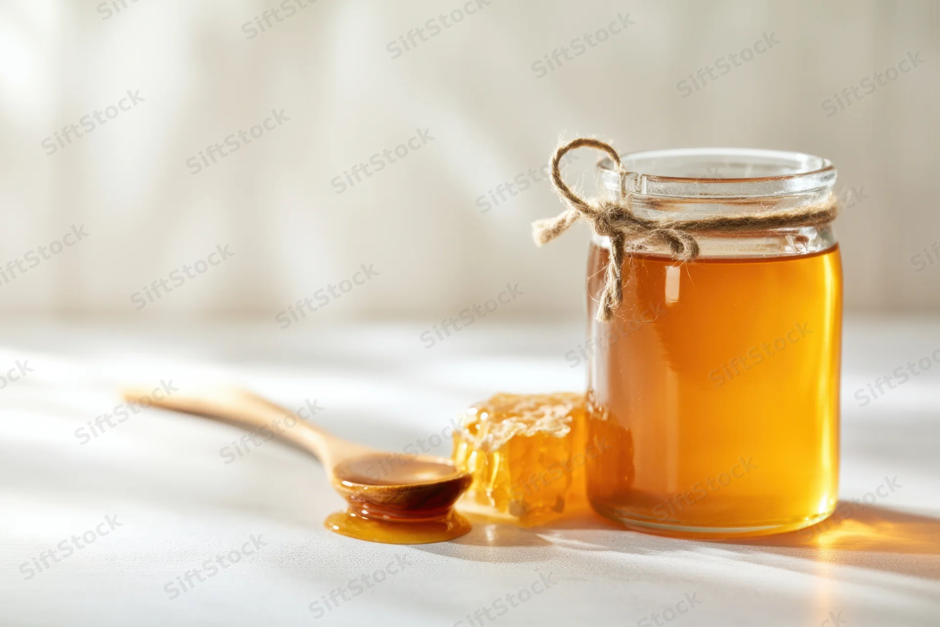 Glass jar of honey with twine loop and wooden spoon beside honeycomb on white table
