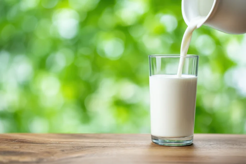 Milk being poured from a ceramic pitcher into a clear cylindrical glass on a wooden table with green bokeh background