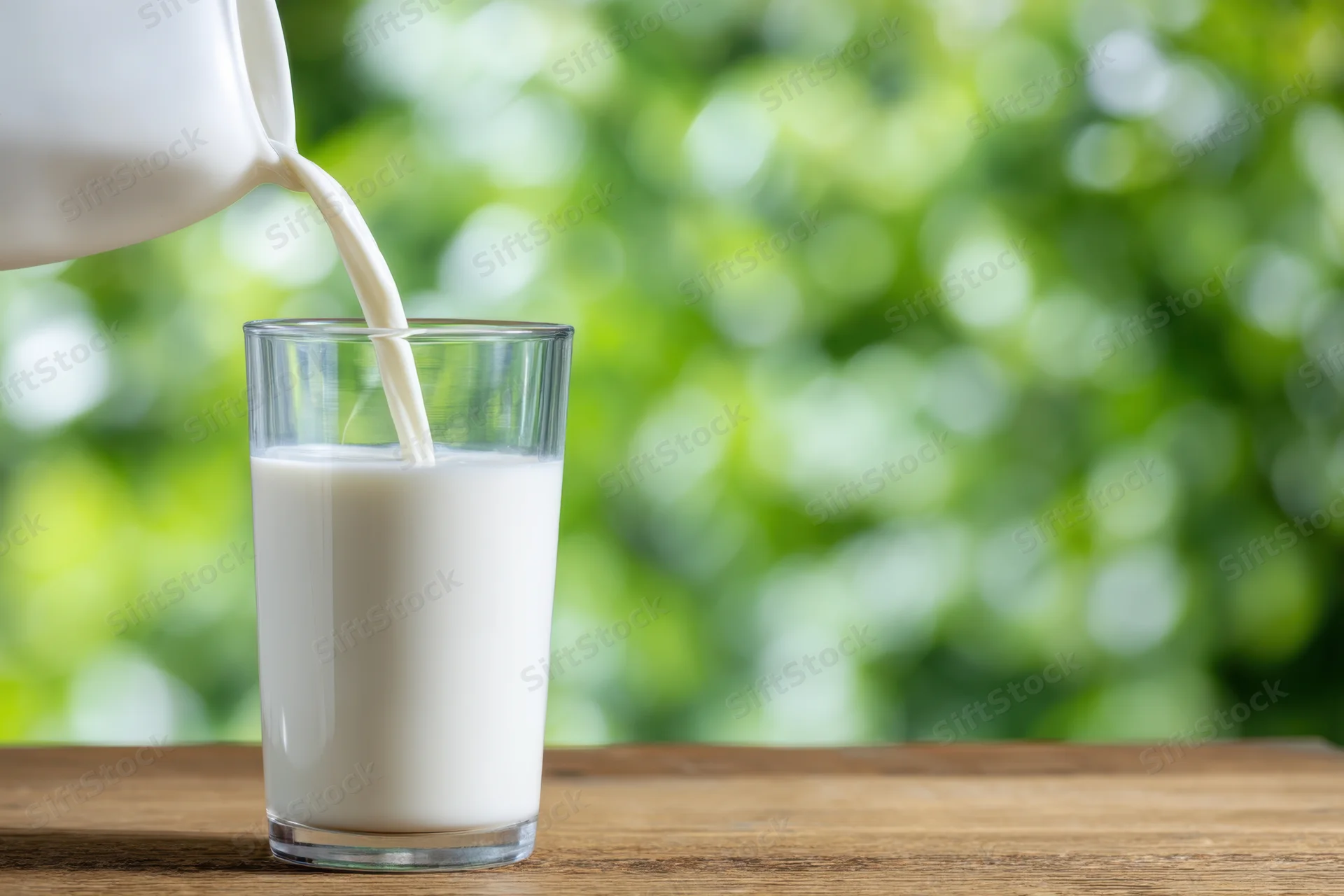 Milk being poured into a clear cylindrical glass on a wooden table with green foliage background