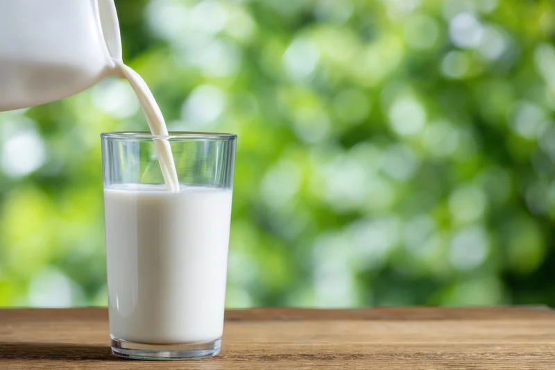 Milk being poured into a clear cylindrical glass on a wooden table with green foliage background