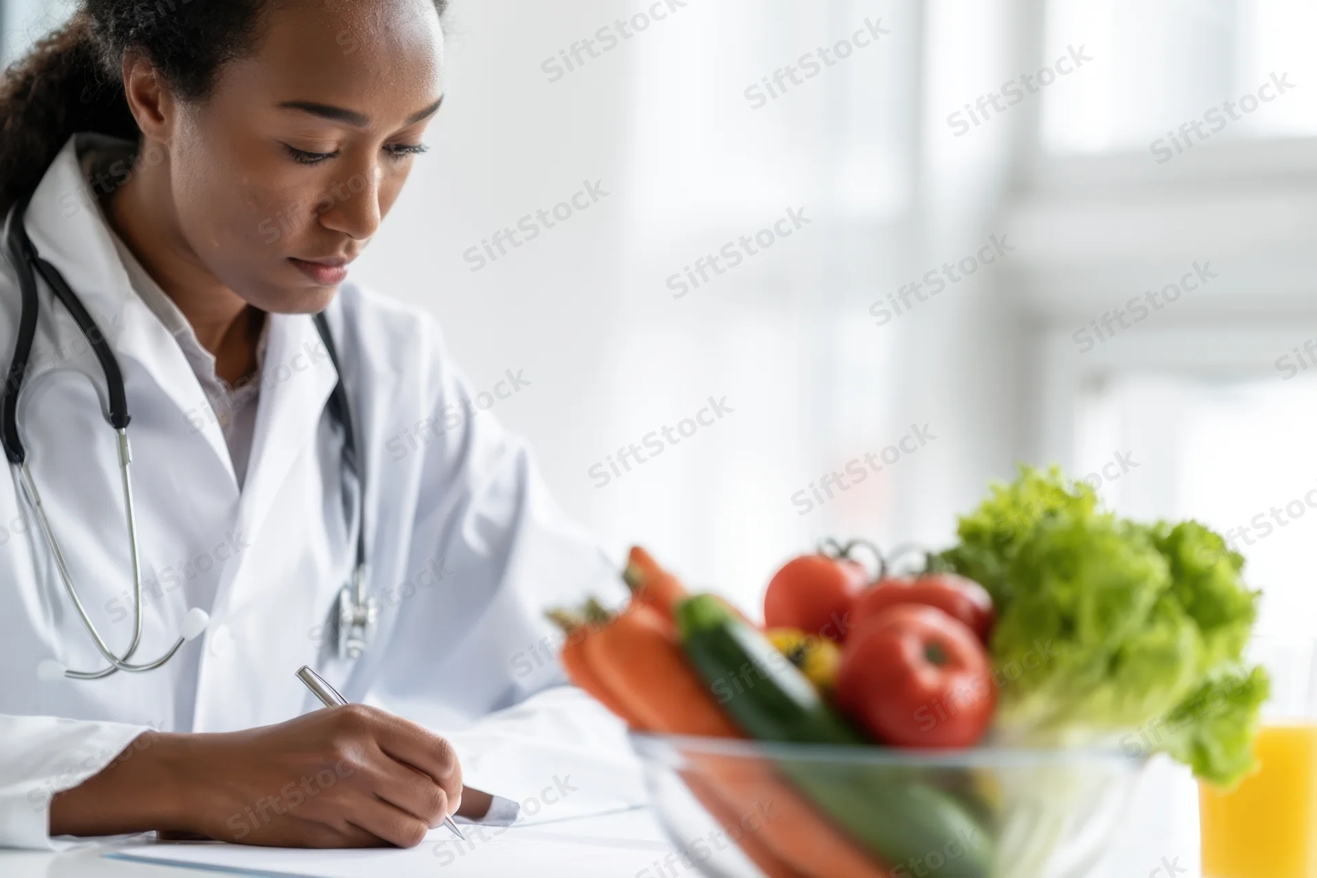 Woman doctor in lab coat writing notes at desk with bowl of fresh vegetables and juice nearby
