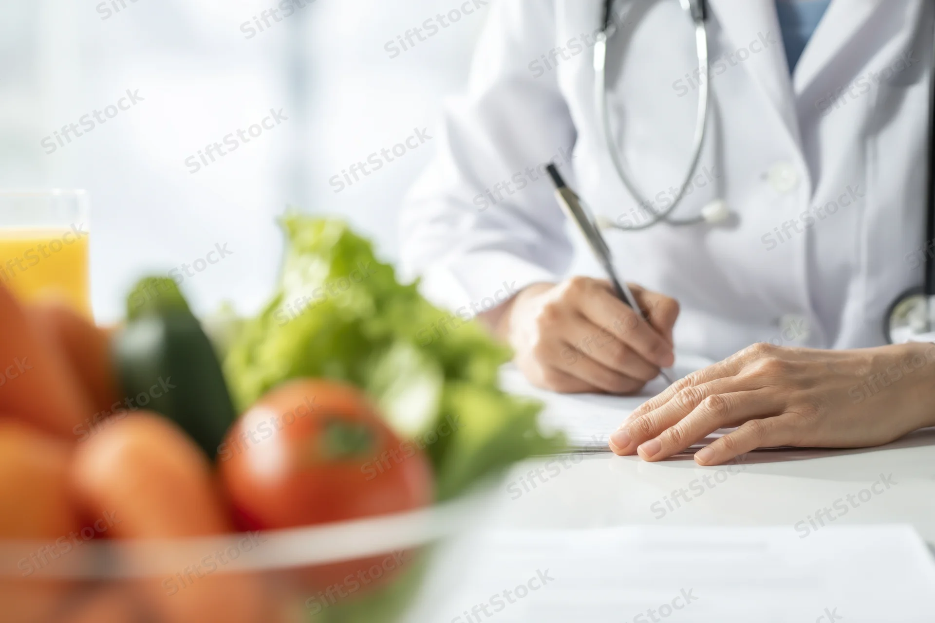 Doctor in white lab coat writing notes at desk with fresh vegetables and juice in foreground