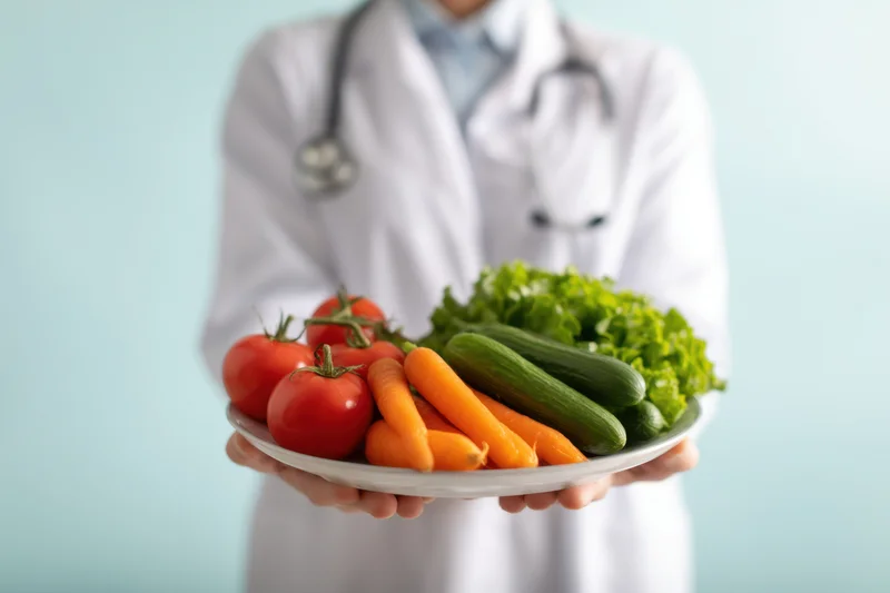 Doctor holding plate of fresh vegetables in front of light blue background