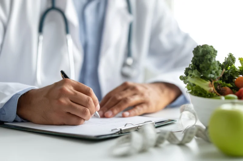 Doctor writing notes on clipboard at desk with vegetables and measuring tape nearby