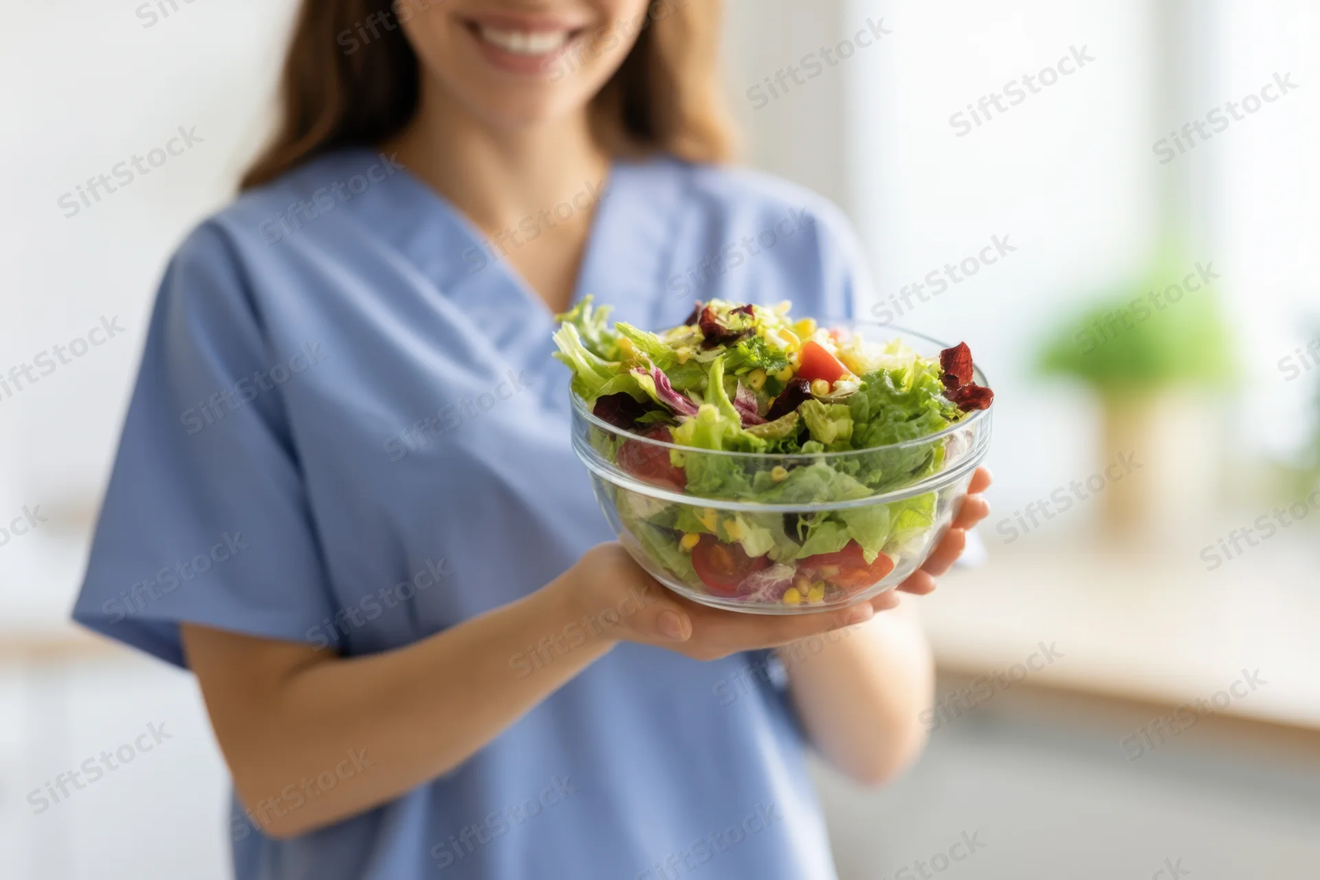 Woman in scrubs holding a glass bowl of fresh salad in a kitchen