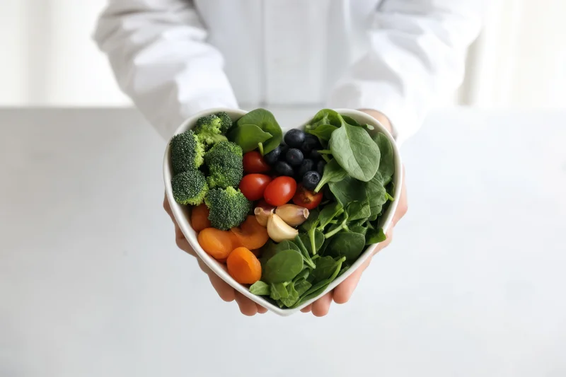 Woman holding heart-shaped bowl filled with fresh vegetables and fruits on white table