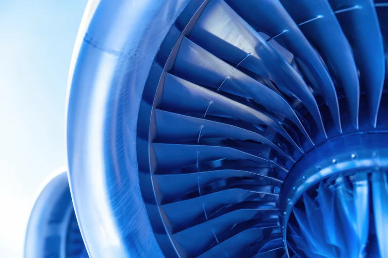 Closeup of jet engine turbine blades showing curved metallic structure in blue tone