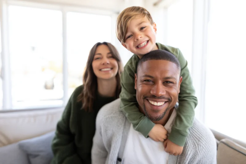 Father carrying son on shoulders while mother sits beside them on couch in bright living room