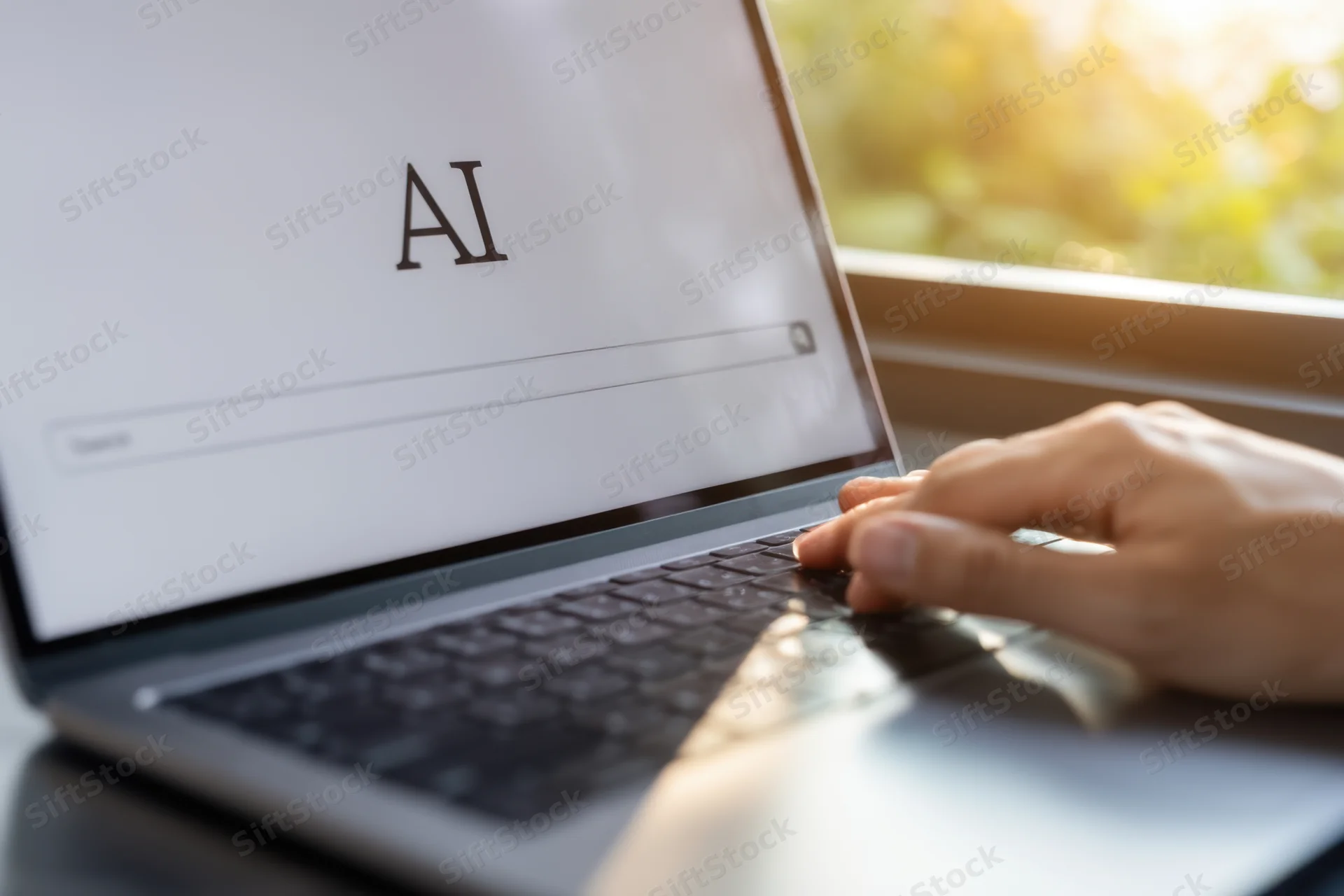 Hand typing on laptop keyboard with ai displayed on screen near window