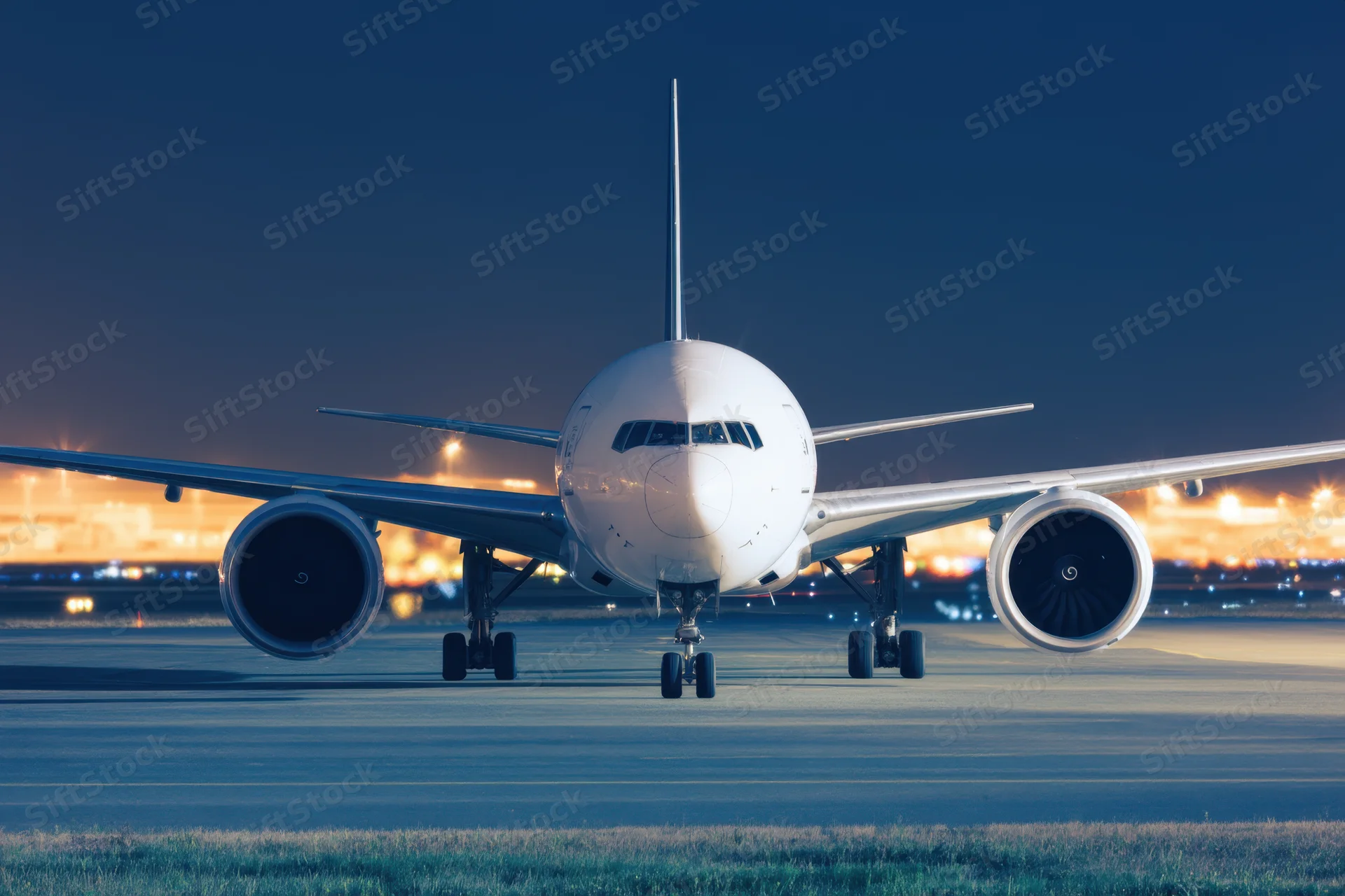 Large commercial airplane viewed head-on on airport tarmac at dusk with city lights in background