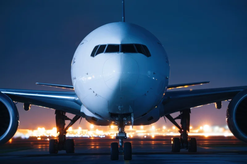 Commercial airplane viewed head-on on tarmac at twilight with airport lights glowing in background