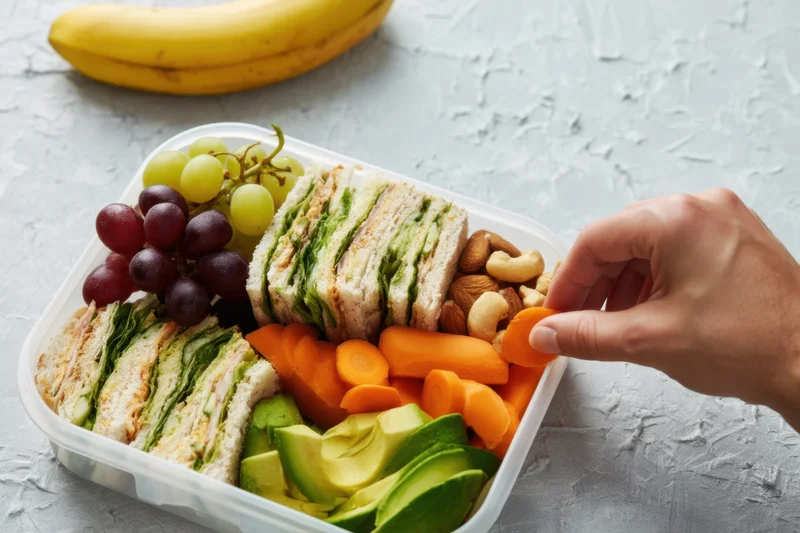 Hand reaching into a lunchbox filled with sandwiches, fruits, vegetables, nuts, and avocado on a textured surface