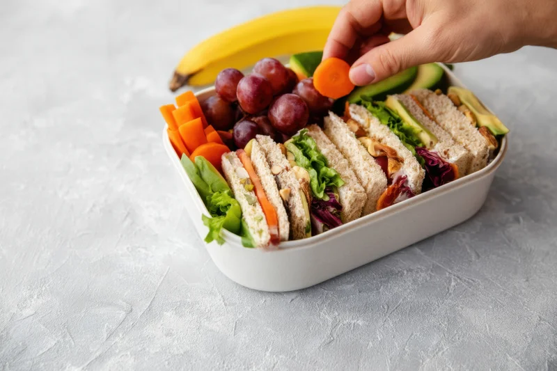 Hand placing a carrot slice into a lunchbox filled with sandwiches, fruits, and vegetables on a gray surface