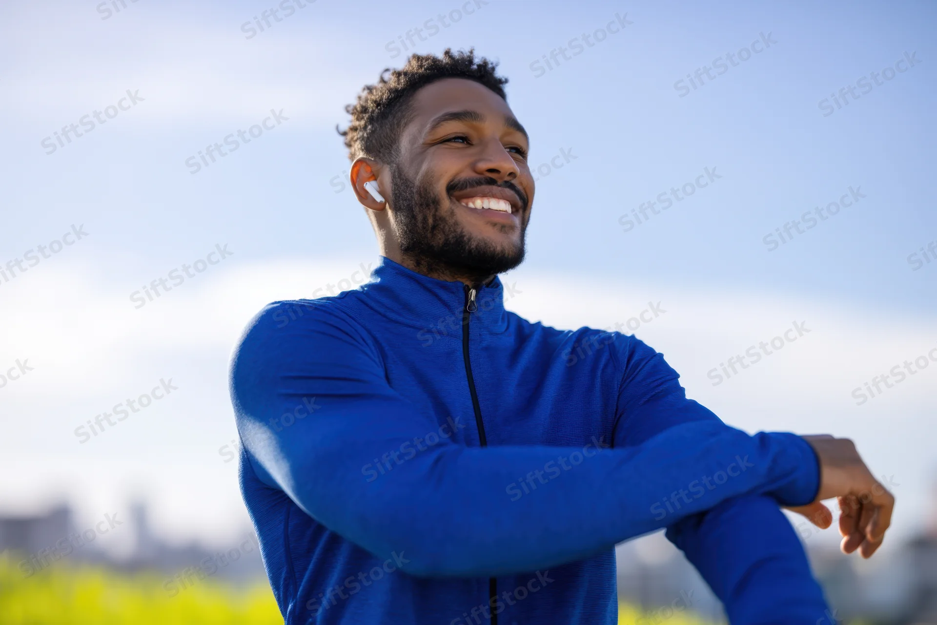 Man stretching outdoors in blue athletic jacket with earbuds smiling under clear sky