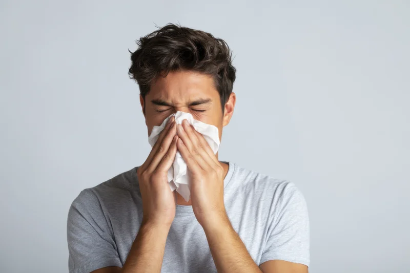 Man holding tissue to his nose while showing signs of illness indoors