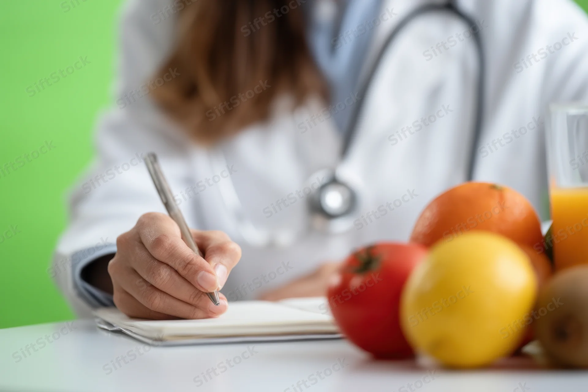 Female doctor in white coat writing in notebook at desk with fresh fruit and juice nearby
