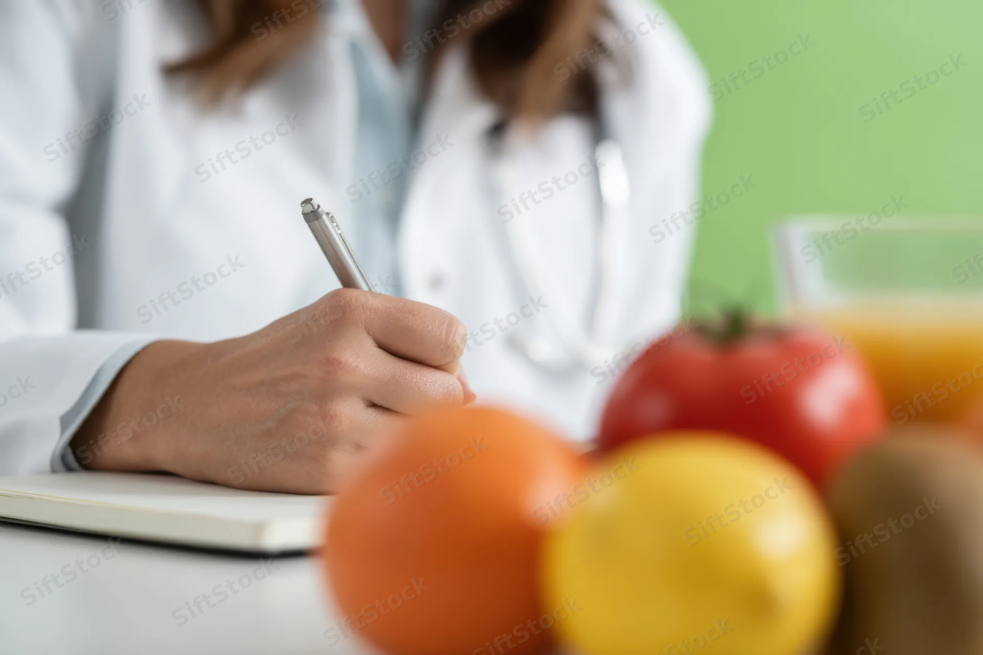 Doctor in white coat writing in notebook with fresh fruits and juice on desk