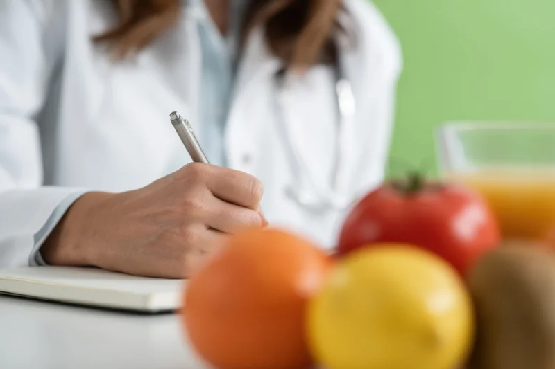 Doctor in white coat writing in notebook with fresh fruits and juice on desk