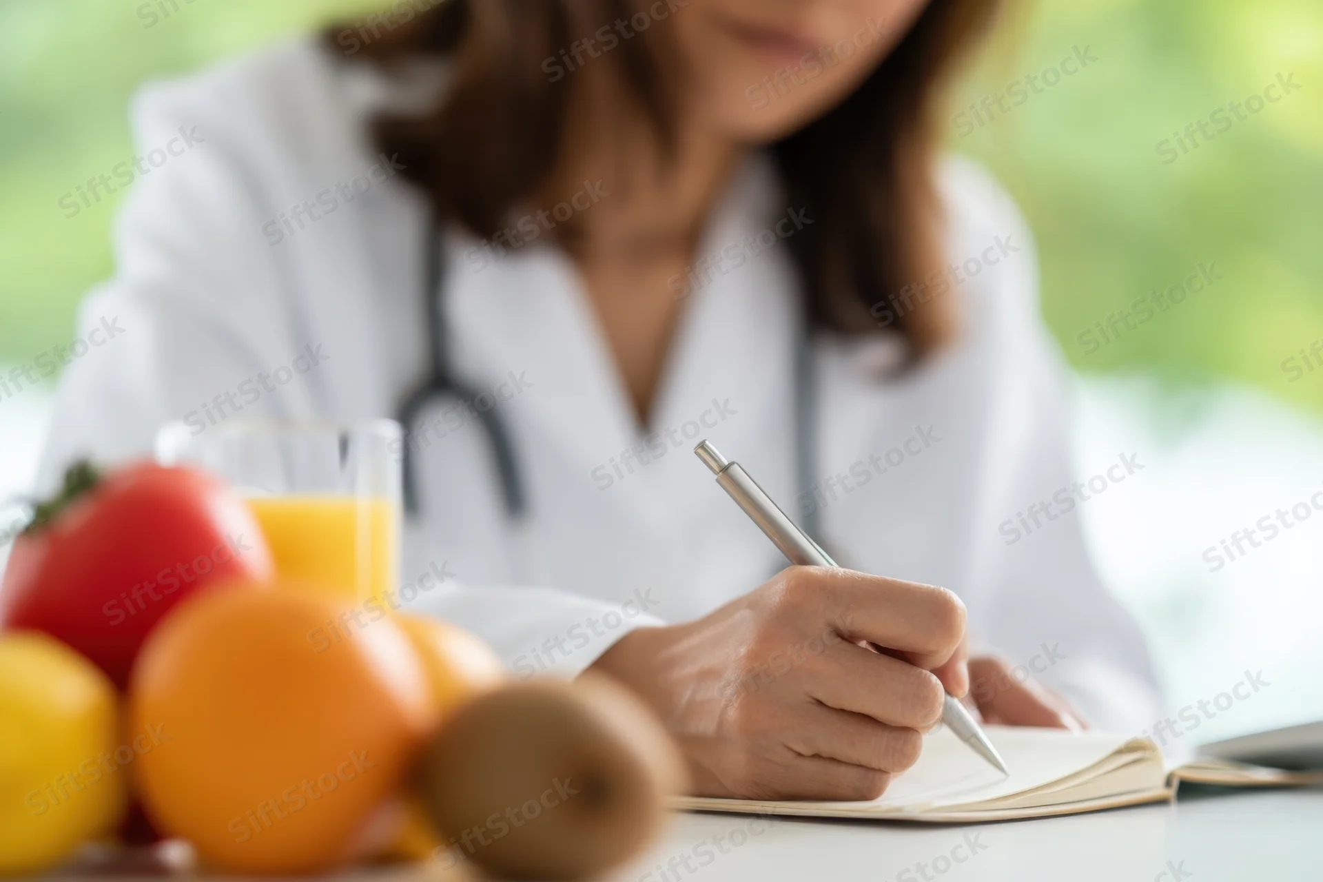 Doctor in white coat writing in notebook at desk with fresh fruit and juice in foreground
