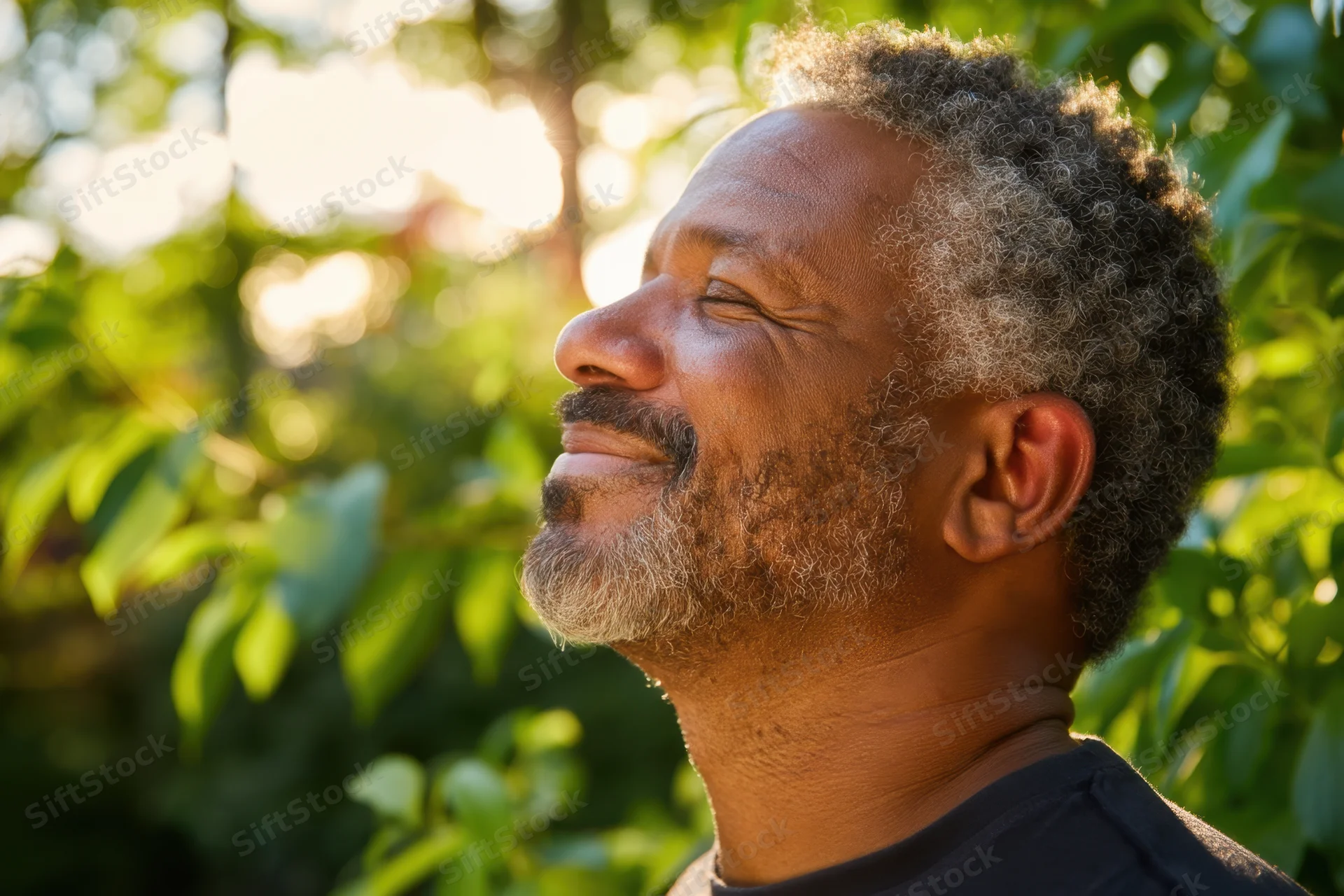 Middle-aged black man smiling in profile outdoors among green foliage in warm sunlight