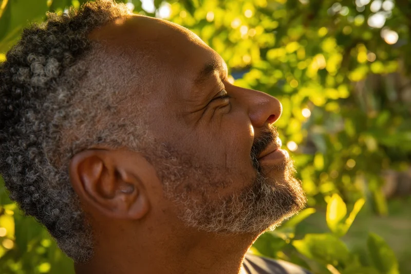 Senior black man with curly gray hair and beard smiling with eyes closed in a sunlit garden surrounded by green foliage