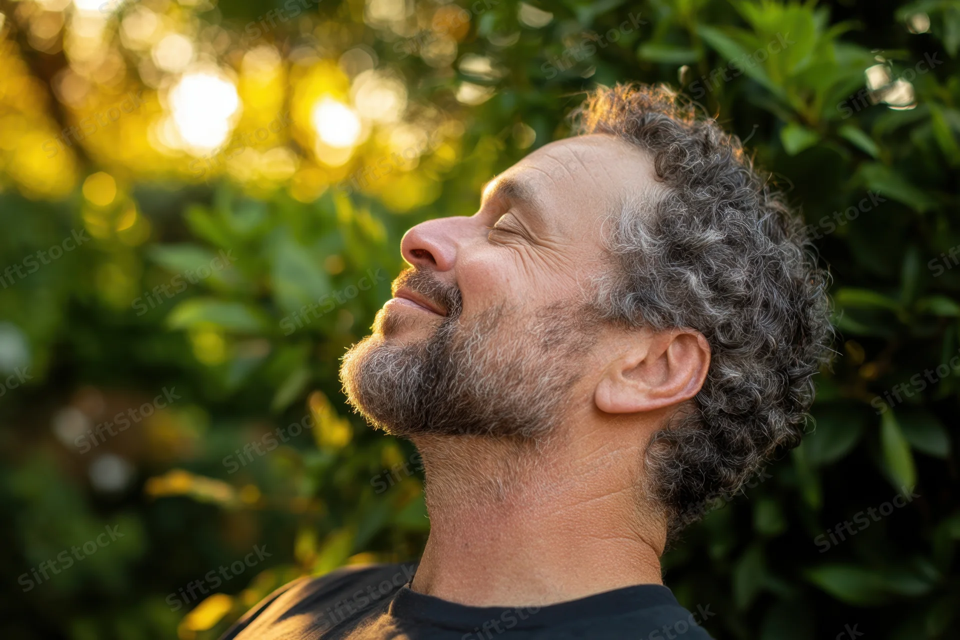 Middle-aged man with curly gray hair and beard breathing deeply with eyes closed in a sunlit garden surrounded by green foliage